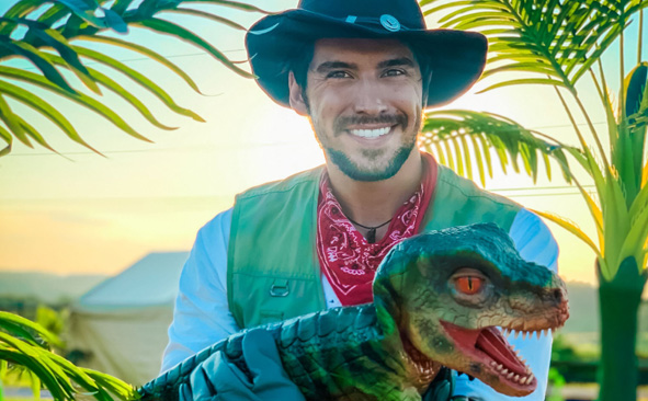 A man in a hat holding a green Tyrannosaurus Rex hand puppet