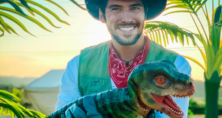 A man in a hat holding a green Tyrannosaurus Rex hand puppet