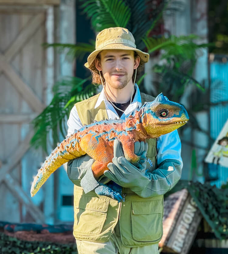 A man in a brown vest holds an orange Carnotaurus hand puppet