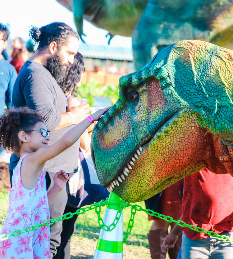 A child in a skirt interacts with a green Tyrannosaurus Rex at Dino Fest