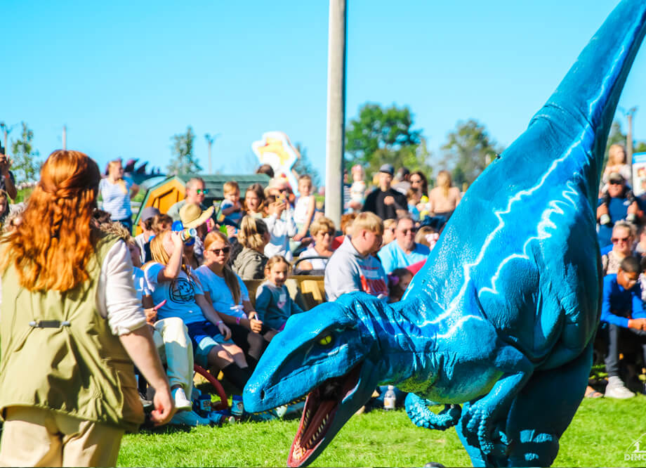 A woman in a brown vest and a blue velociraptor perform at the center of an event at Jurassic Farm, surrounded by many spectators