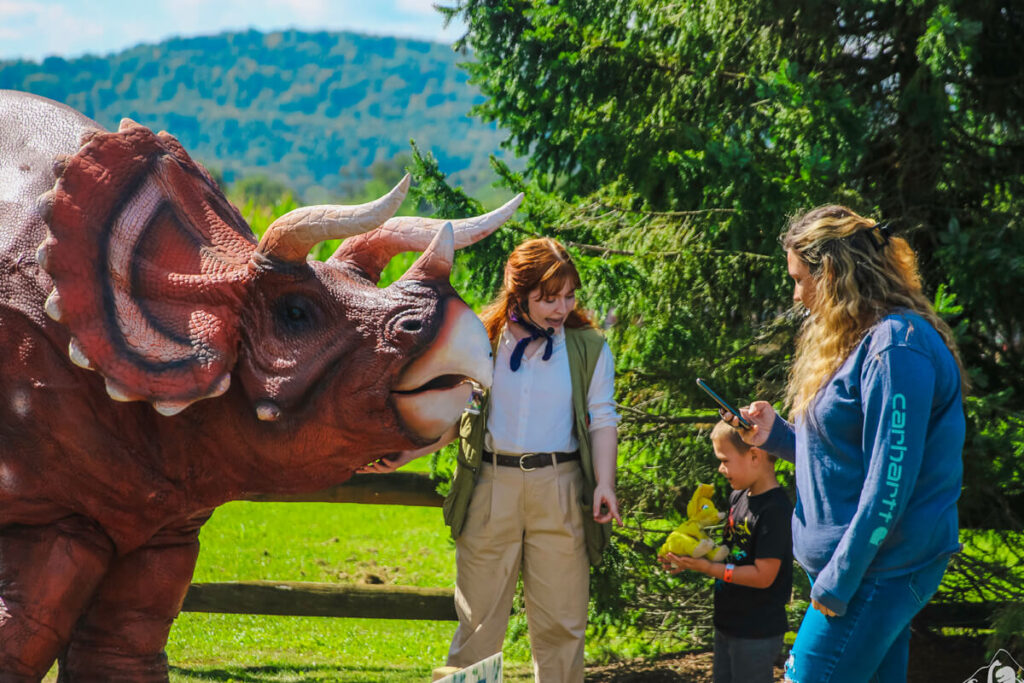 A woman wearing a brown vest and with reddish-brown hair, holding an orange triceratops, interacts with a child at Dinofest