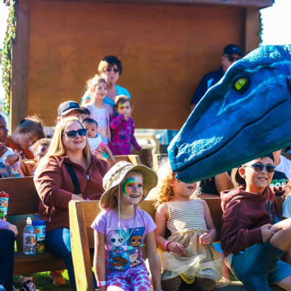 A blue 2-meter-tall velociraptor interacts with a child in a pink dress at the Jurassic Farm