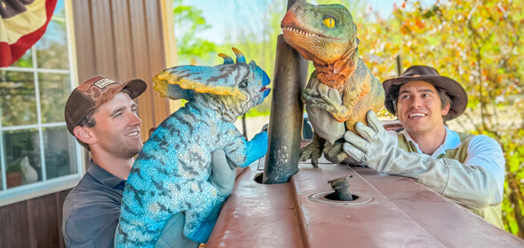 Two men took a photo at DinoFest holding a blue Triceratops and a green Tyrannosaurus Rex puppet respectively