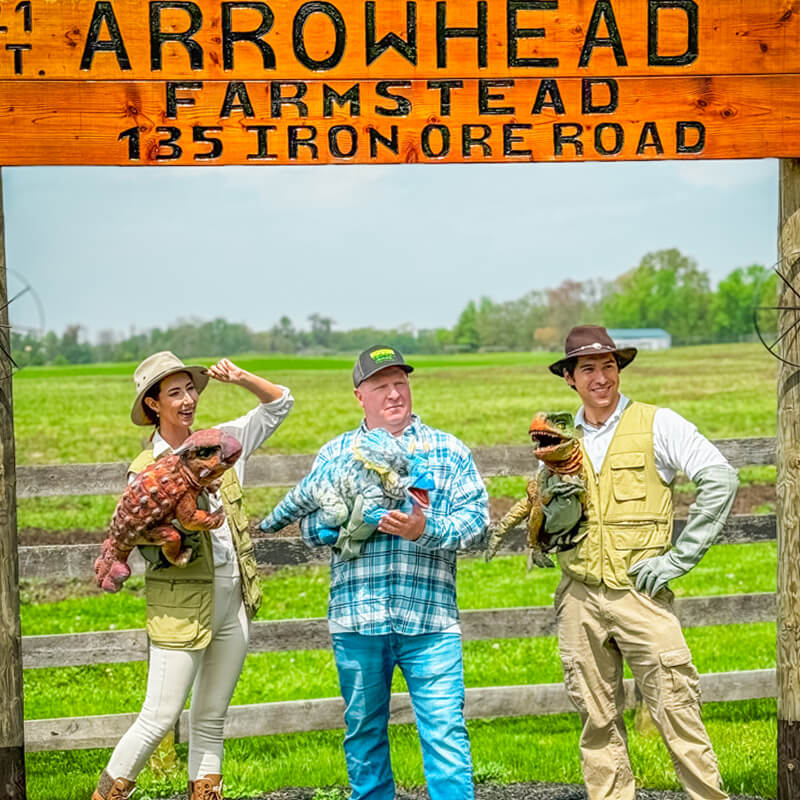 A man in a brown vest, a woman in a brown vest, and a man in a blue and white plaid shirt, holding an Ankylosaurus, a Triceratops, and a Tyrannosaurus Rex puppets, took a photo at the Jurassic Farm