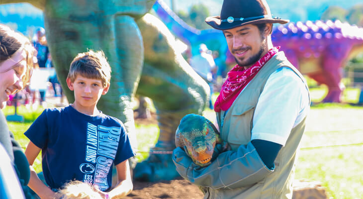 A man in an apricot vest is holding a green Tyrannosaurus Rex puppet and interacting with a little girl.