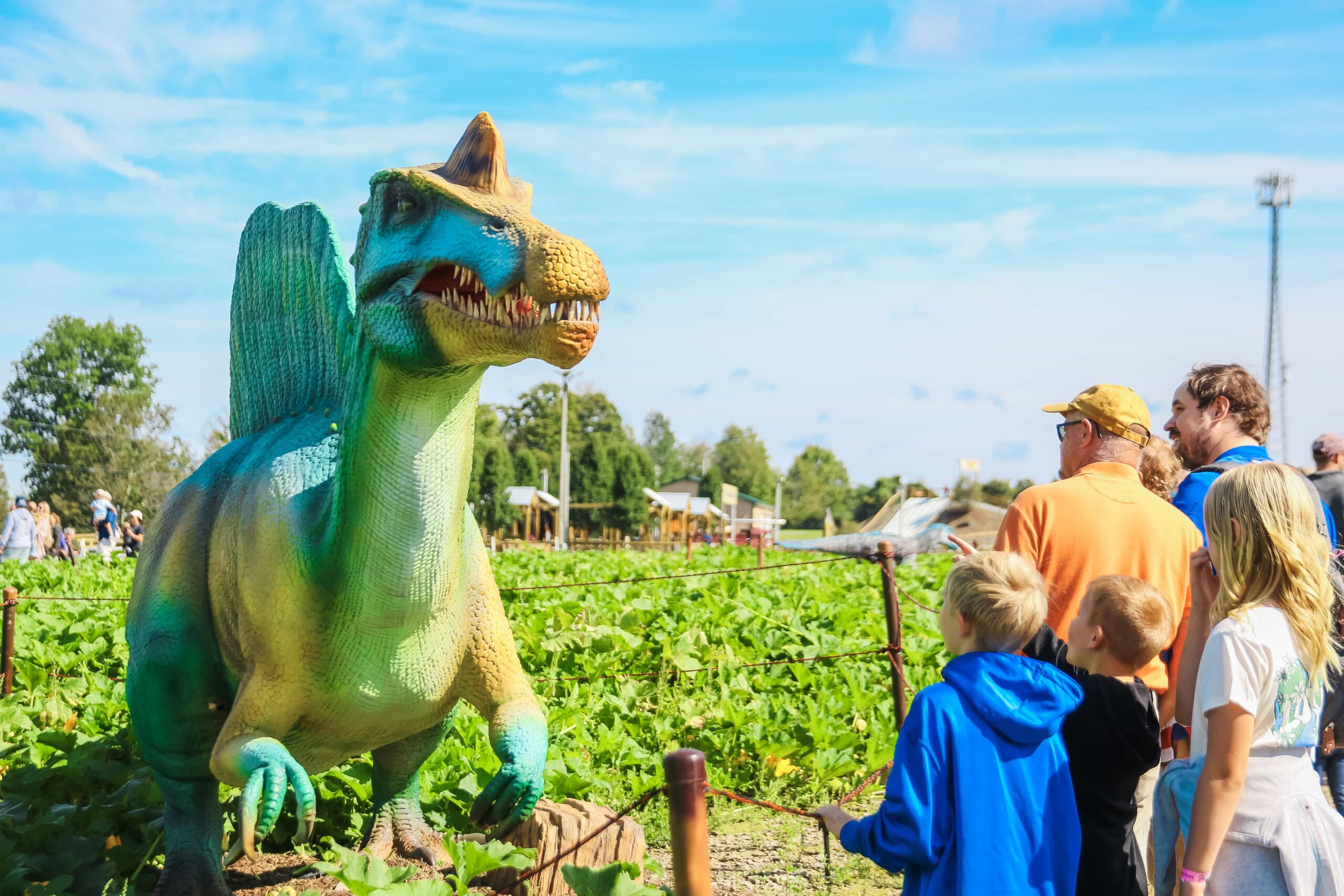 Two men and three children look at a green animatronic Spinosaurus in a vegetable patch at Dino Days