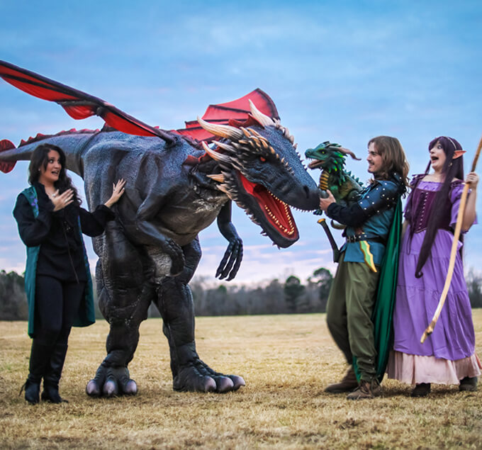 A woman in a purple dress, a woman in a blue dress, a man in armor, and a flying dragon with red and black skin interacting on a lawn