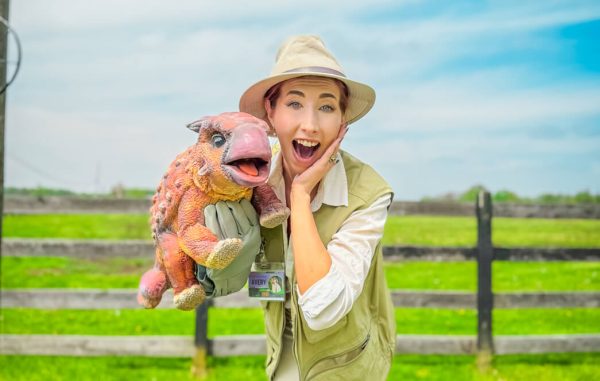 A woman in an apricot vest holding an orange Ankylosaurus hand puppet